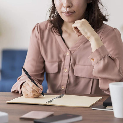 Woman in a pink shirt writing in a notebook at a desk with a cup of coffee.